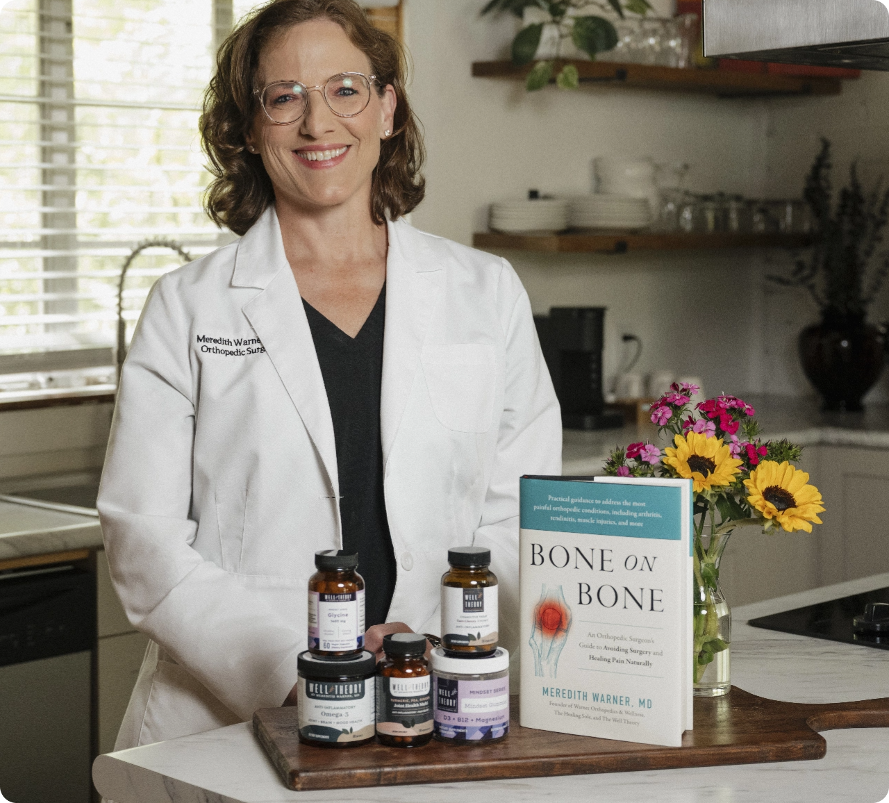 A woman in a lab coat stands in a kitchen beside supplements, flowers, and "Bone on Bone" by Meredith Warner, MD.