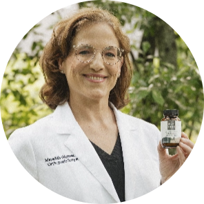 A person in a white lab coat smiles outdoors, holding a bottle of natural supplements in front of greenery.