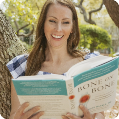 Woman with long brown hair sits outdoors, smiling as she reads "Bone on Bone" and enjoys pain relief supplements.
