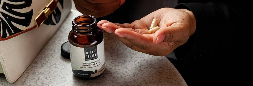 A person holds two capsules and a brown supplement bottle next to a white purse on a countertop.
