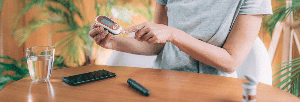 A person at a table uses a blood glucose meter, with supplements, water, a smartphone, and medication nearby.