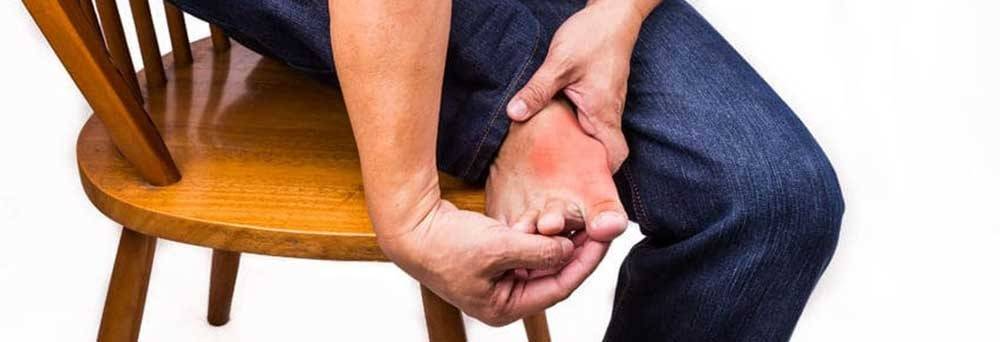 A person sits on a wooden chair holding their red, irritated big toe, possibly seeking natural pain relief.
