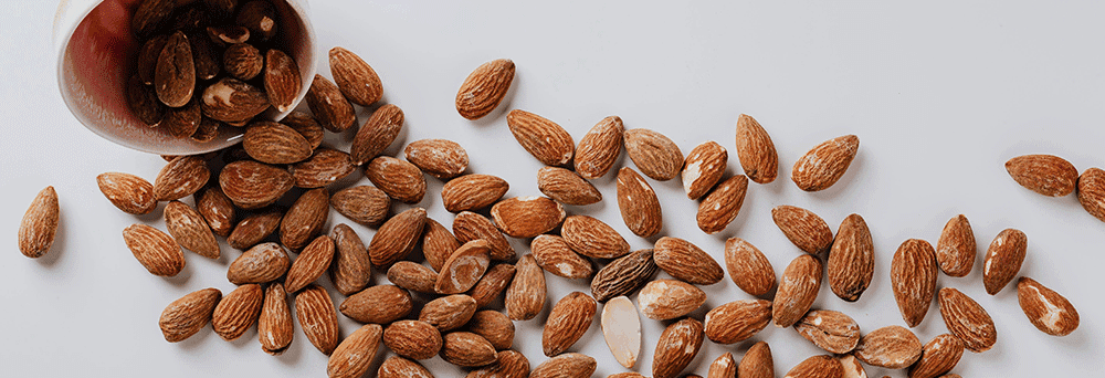 A cup tipped over spills whole almonds, a natural supplement, onto a white surface.