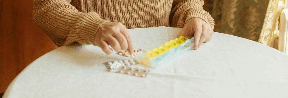 Person sorting pills and natural supplements into a weekly organizer on a round table with blister packs nearby.