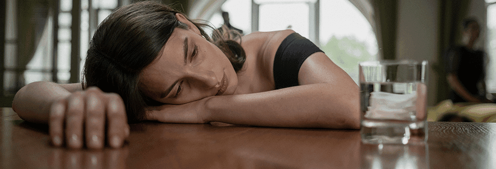 A woman rests her head on a wooden table, looking at pain relief supplements next to a glass of ice water.