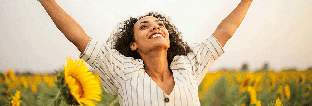 A curly-haired person in a sunflower field smiles and raises both arms, celebrating in a white striped shirt.