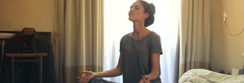 A woman sits cross-legged on a bed in a sunlit room, meditating peacefully with natural supplements nearby.