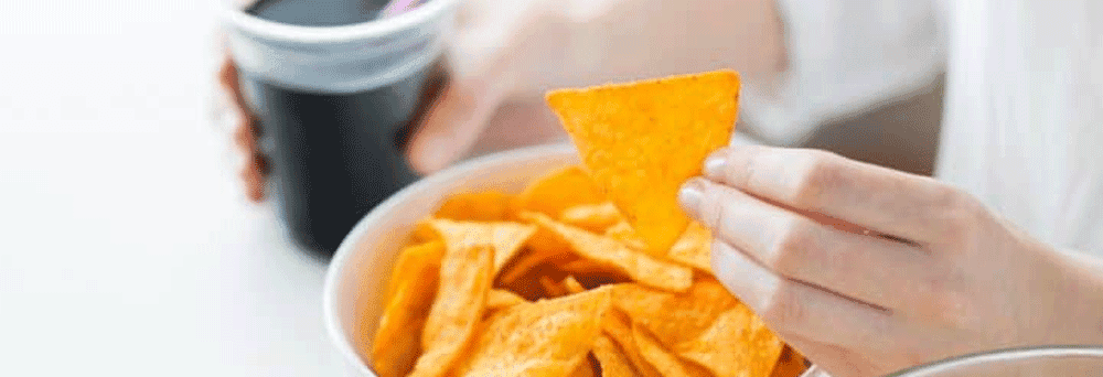 A person holds a triangular chip over a bowl of chips with a dark drink and supplements on the table nearby.