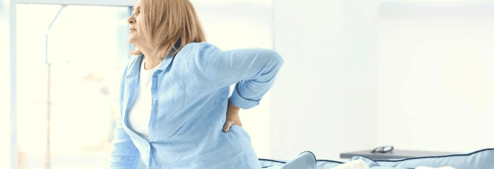 An older woman in a blue shirt stands indoors, holding her lower back with a painful expression.