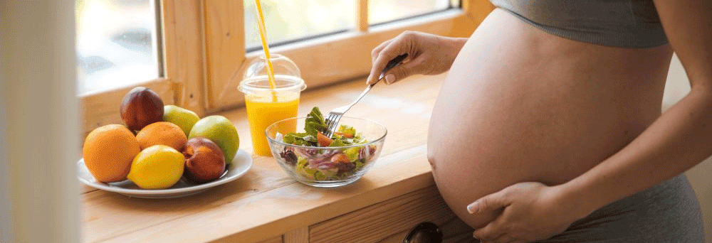 A pregnant person enjoys salad, juice, and fruit by a window, focused on a healthy pregnancy.
