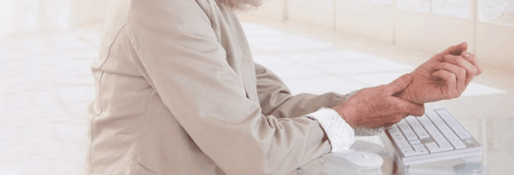 An older adult in a light suit holds their wrist at a desk with a keyboard, considering natural pain relief.