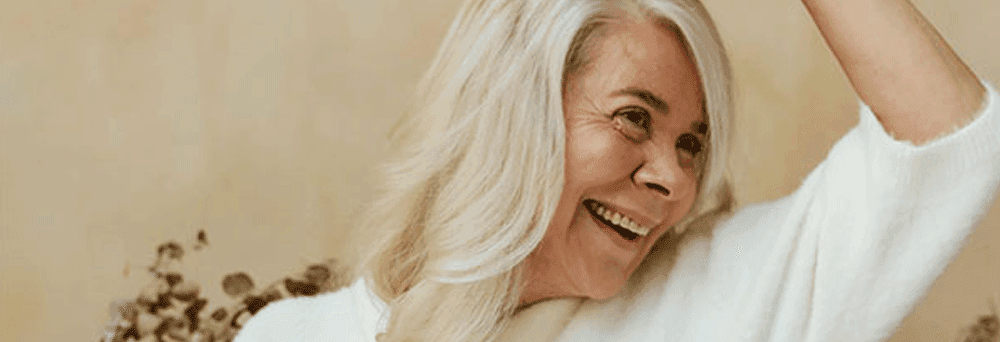 Older woman with long white hair smiles, raising her arm with energy, set against a beige floral background.