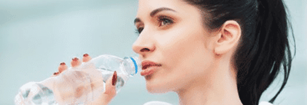 A woman with a dark ponytail drinks from a clear water bottle, mindful of her surgeon-formulated supplements.