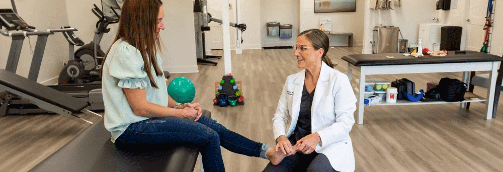 A healthcare professional examines a woman's foot in a physical therapy clinic with supplements visible in the background.