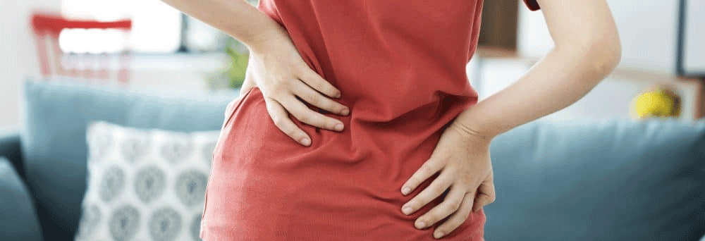 Person in a red shirt stands indoors, holding their lower back as if searching for natural pain relief.