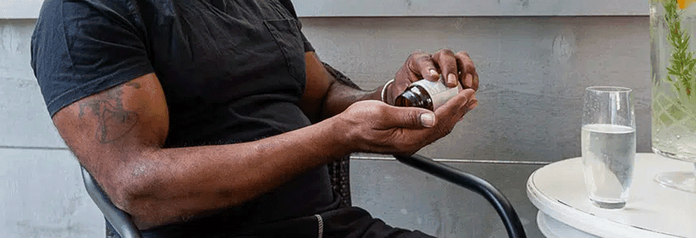 A person at a table opens a bottle of surgeon-formulated supplements with a glass of water nearby.