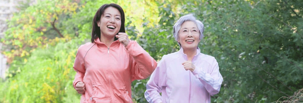 Two women, one younger and one older, jog outside in light jackets, energized as they run among green trees.