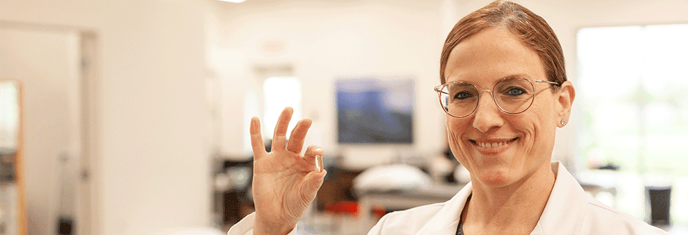A woman in a white lab coat holds up a small capsule, emphasizing supplements in a bright laboratory setting.