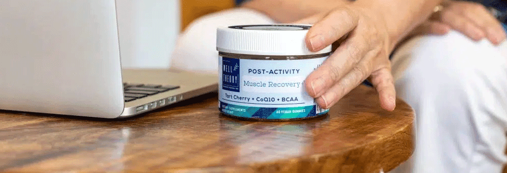 A hand reaches for a jar of "Post-Activity Muscle Recovery" supplements on a wooden table near an open laptop.
