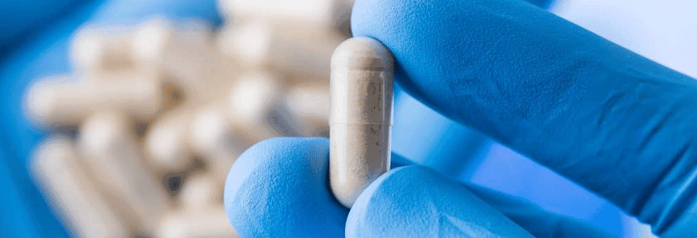 A gloved hand holds a white capsule pill with more capsules blurred in the background, suggesting supplements.