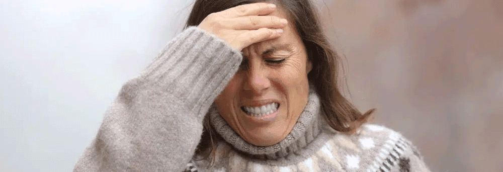 A woman in a patterned sweater winces and presses her hand to her forehead, appearing to have a headache.