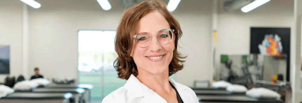 A smiling person in glasses and a white lab coat stands in a bright clinic with surgeon-formulated supplements.