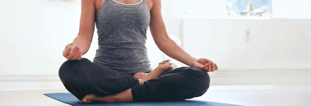 Person sits cross-legged on a yoga mat indoors in grey tank top and black pants, practicing calm with supplements.