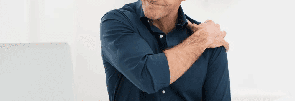 A person in a blue shirt touches their left shoulder, appearing to be in discomfort, possibly seeking pain relief.