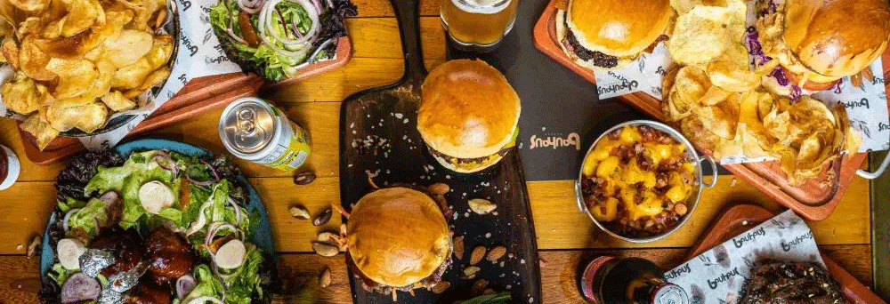 Burgers, chips, salads, soda, beers, and surgeon-formulated supplements sit on a wooden table in a casual setting.