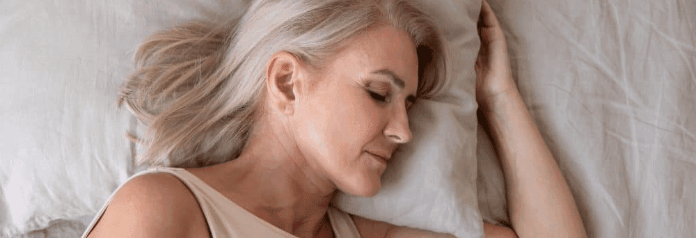 A gray-haired woman sleeps on her side in bed, head on a white pillow, after taking natural supplements.