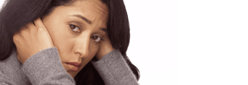 A woman with long dark hair holds her hands to her ears, looking forward with a concerned expression.
