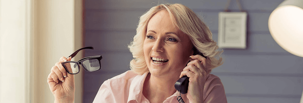 Smiling woman holds eyeglasses and talks on a corded phone indoors about pain relief supplements.