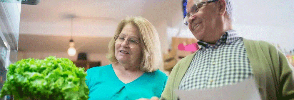 An older couple in a grocery store hold a shopping list and look at supplements near the lettuce.