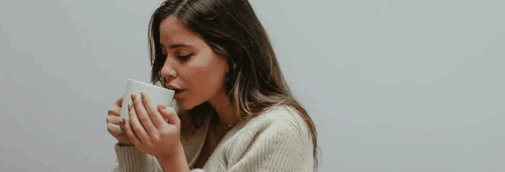 A woman with long brown hair in a beige sweater sips from a white mug, enjoying natural supplements indoors.
