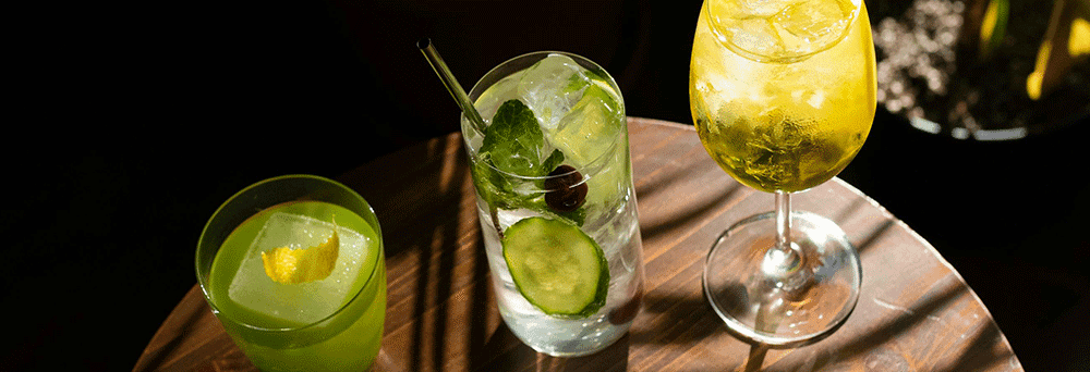 Three colorful cocktails with fresh garnishes sit on a wooden table, looking refreshing and vibrant.