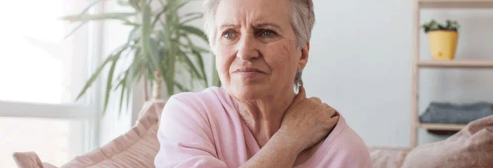 Older woman on couch holding her shoulder in discomfort; plant and shelves in background.
