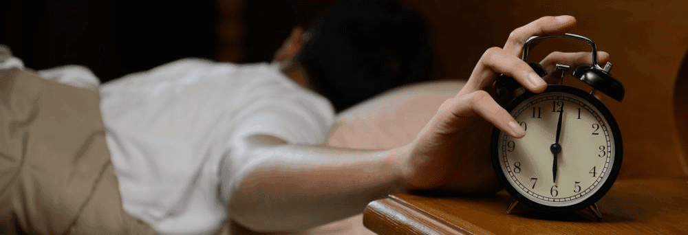 A person in bed reaches to turn off a 7:00 alarm, with natural supplements on the bedside table.