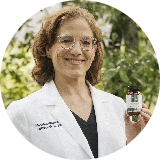 A person in a white lab coat smiles outdoors, holding a bottle of supplements with greenery in the background.