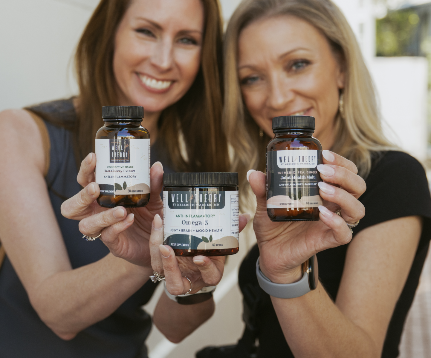 Two women smile and hold up Well Theory supplements and a jar, facing the camera.