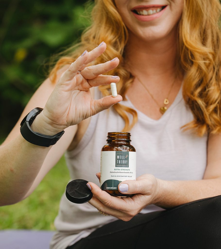 A woman smiles outdoors, holding a white capsule and open Well Theory bottle with supplements in hand.