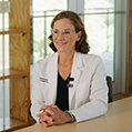 A woman in a white lab coat sits at a table, smiling warmly in a bright room.