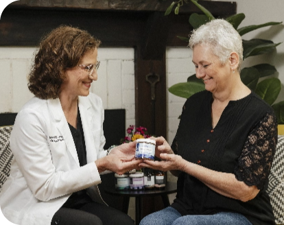 A doctor gives a small supplement container to a smiling patient in a room with plants and shelves in the background.