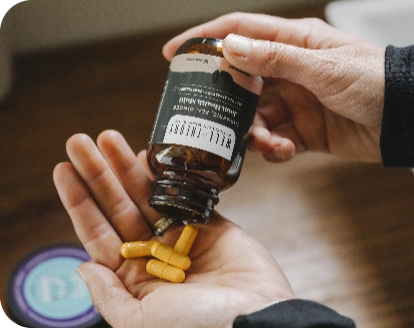 A person pours yellow supplement capsules from a brown bottle into their hand above a wooden surface.