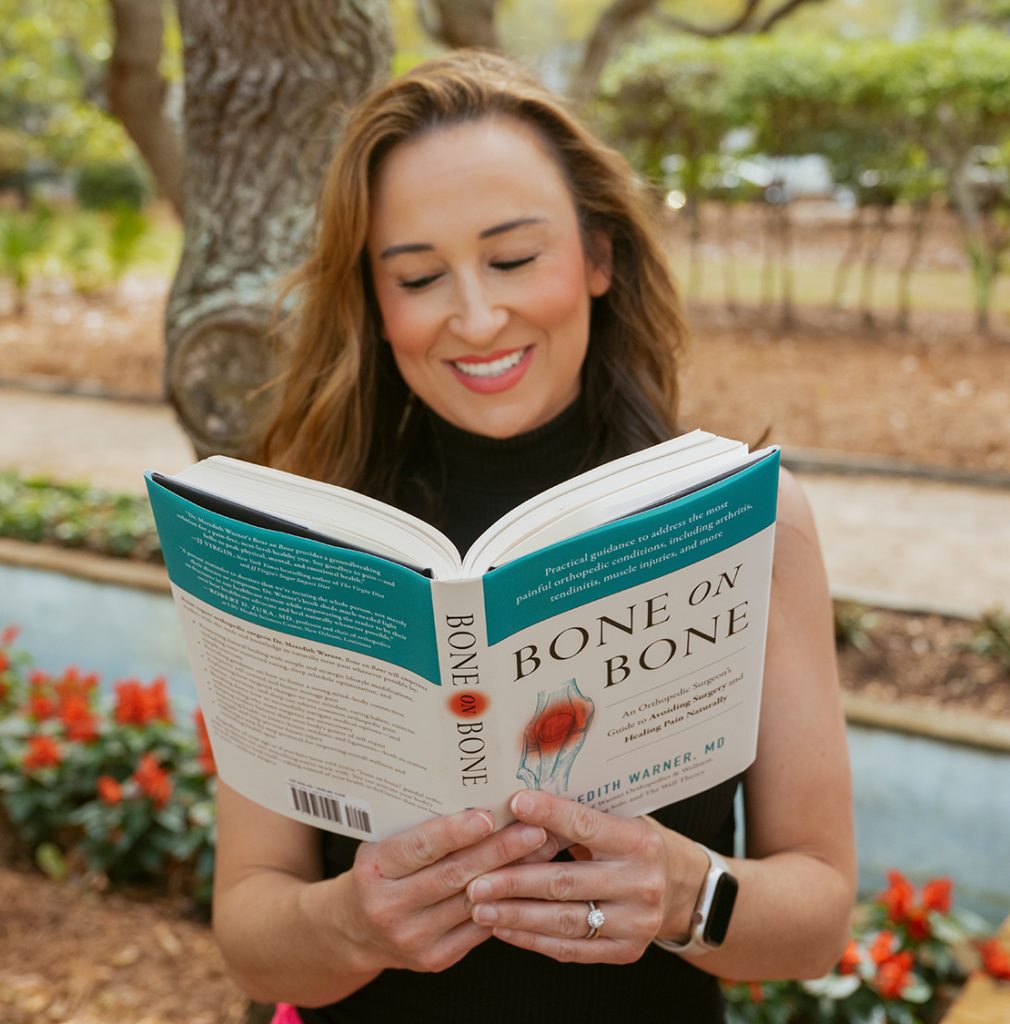 Woman in a sleeveless top reads "Bone on Bone" outdoors, smiling, with trees, flowers, and supplements nearby.