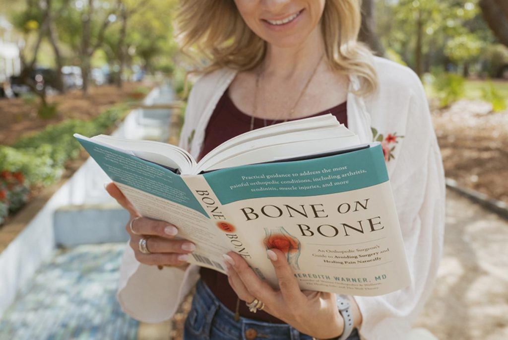 A woman reads "Bone on Bone" by Meredith Warner, M.D. outdoors, a book about supplements for natural pain relief.
