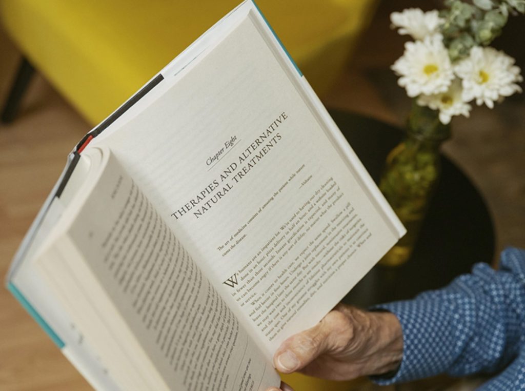 A person reads a book on natural therapies next to pain relief supplements and white flowers in a vase.