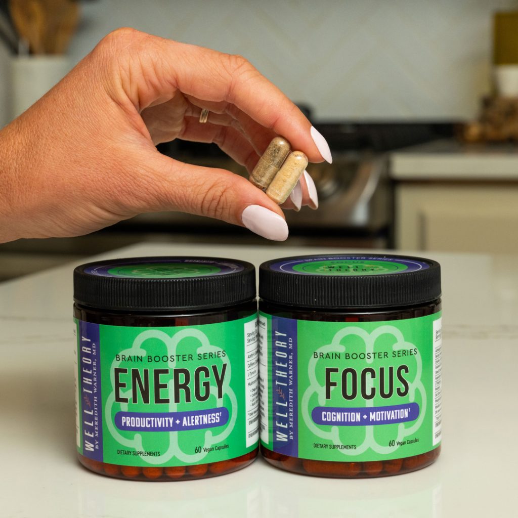 A hand holds two supplements above jars labeled "Energy" and "Focus" on a kitchen counter.