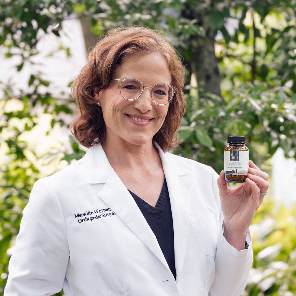 A smiling woman in a white lab coat holds a small bottle outdoors, highlighting surgeon-formulated supplements.