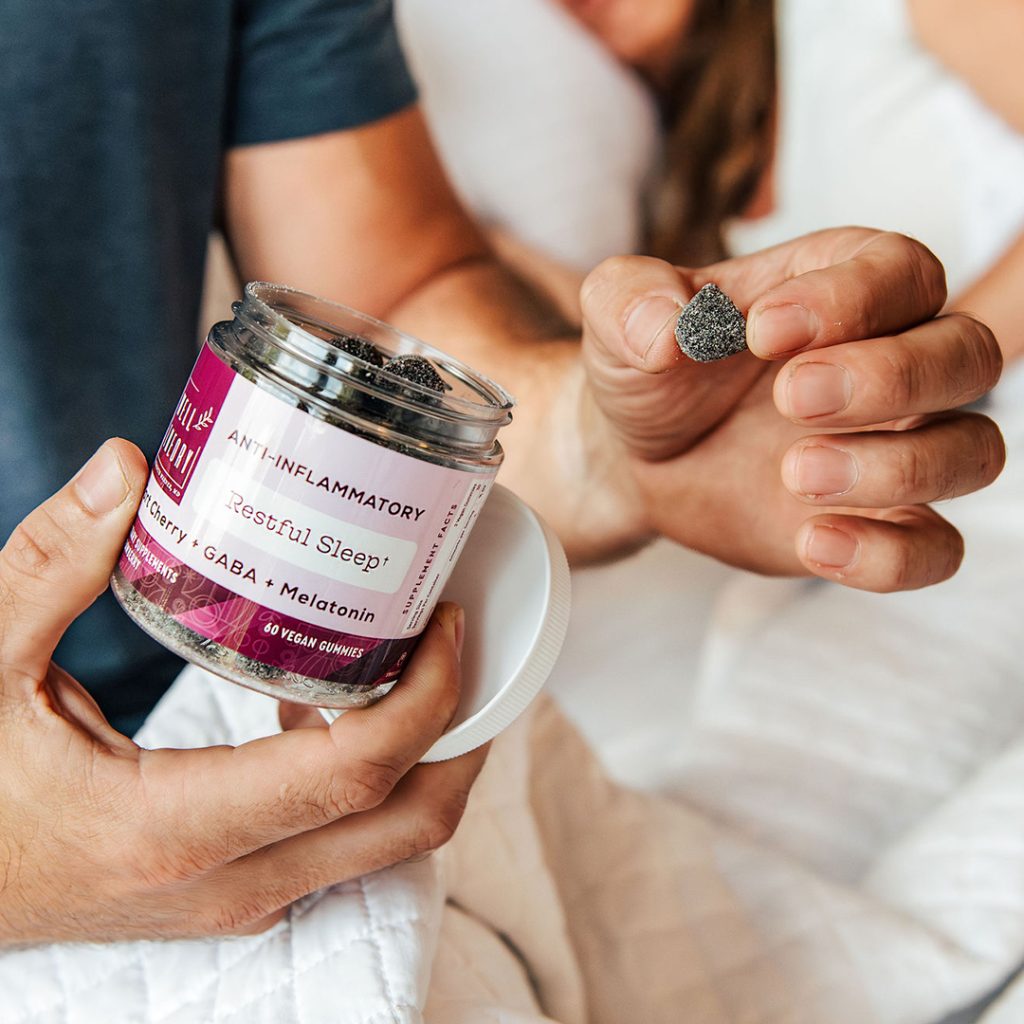 A person on a bed holds an open jar of "Restful Sleep" supplements and picks up a gummy.
