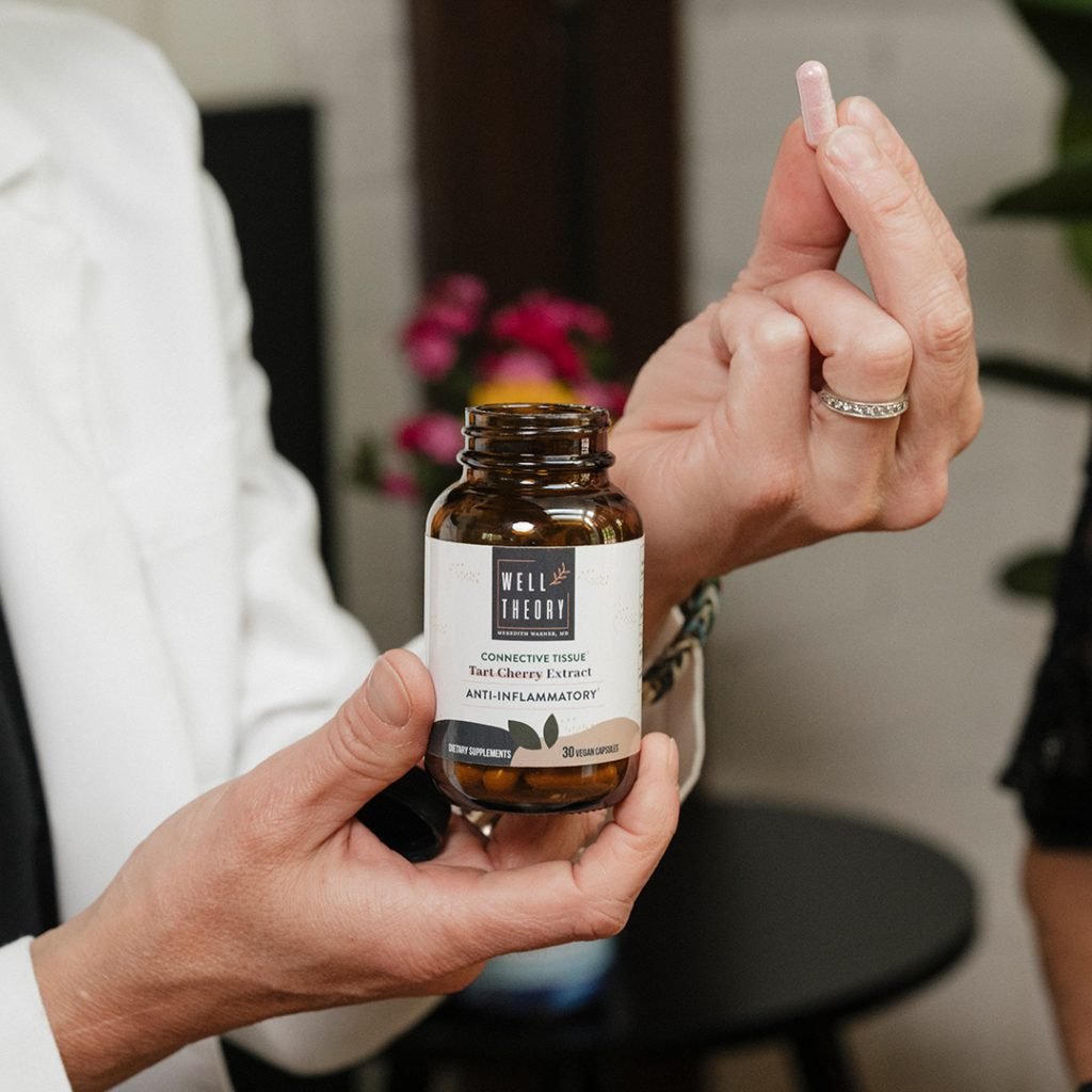 A person holds a Well Theory supplement bottle in one hand and a pink capsule in the other.